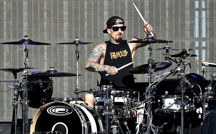 Travis Barker mengenakan kacamata hitam, topi hitam, dan tanktop hitam sedang bermain drum